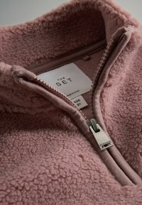 Fleecetröja i mjuk rosa med hög krage, halvdäck och texturerad yta. Lappen inuti säger "THE SET," storlek 6Y.
