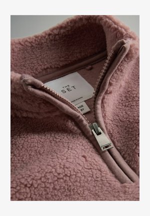 Fleecani pulover u mekom rozoj boji s visokim ovratnikom, polu-zipperom i teksturiranom površinom. Unutrašnja oznaka glasi "THE SET", veličina 6Y.