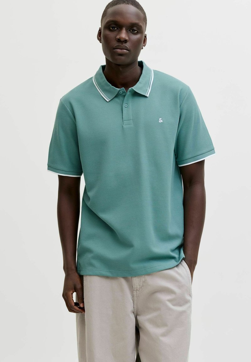 Jaunas vyras dėvi žalsvą trumpomis rankovėmis polo marškinėlį su baltu apvadu ir šviesiai smėlio spalvos kelnes, stovintis prieš šviesią fono sieną.