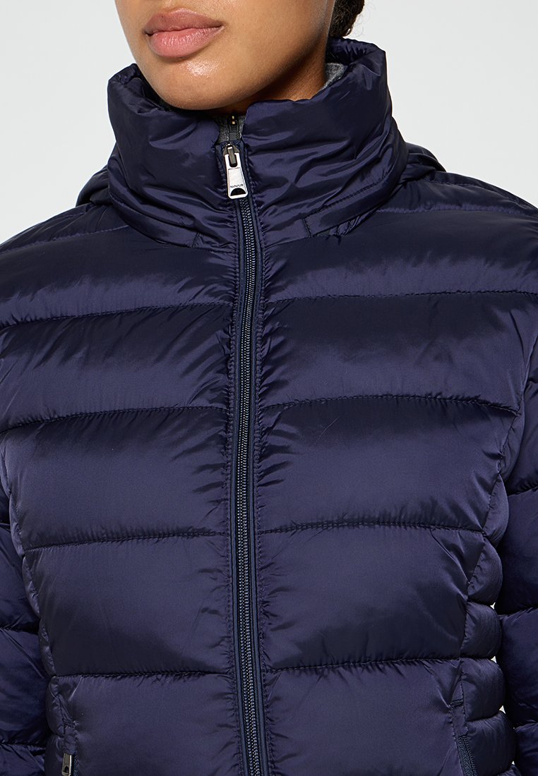 Navy pufferjack met een hoge kraag, horizontaal gestikt patroon en een rits aan de voorkant. Materiaal lijkt glanzend met een gladde textuur.