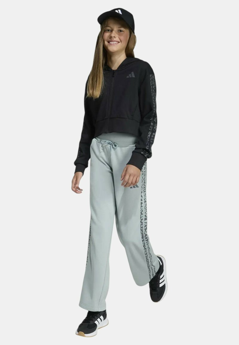 Fille souriante portant un sweat à capuche Adidas noir, un pantalon de survêtement Adidas gris clair, des baskets Adidas noires et une casquette Adidas noire, debout avec assurance.