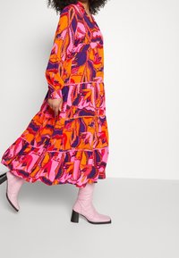 Lange, gelaagde jurk met een levendige bloemenprint in oranje, roze en paarse tinten, met lange mouwen en een ronde halslijn.