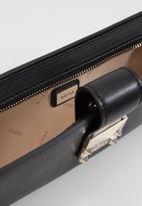 Mustat nahkalompakko, jossa on kultainen vetoketju, merkkilogo sisävuorauksessa ja koristeellinen niittikoriste soljessa.