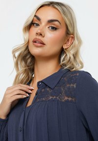 Navy blue button-up blouse with lace detailing on the shoulders, featuring a collared neck and textured fabric. Long sleeves with gathered cuffs.