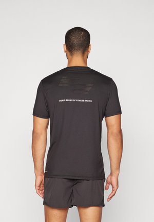 Homme debout, de dos, portant un t-shirt noir avec le texte "World Series of Fitness Racing" et un short noir sur un fond uni.