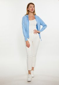 Felpa zip con capucha azzurra chiara con una finitura testurizzata, indossata sopra un top bianco e abbinata a pantaloni bianchi attillati e sneakers bianche.