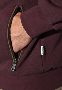 Burgunderfarbene Kapuzenjacke mit Reißverschluss aus strukturiertem Stoff, seitlicher Tasche mit Metallreißverschluss und kleinem weißen Etikett am unteren Saum.