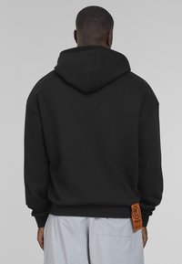 Black hoodie with a large, soft hood and a rectangular orange tag showing the brand on the lower side, made of textured fabric.