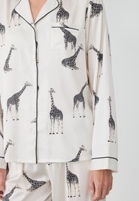 Jedwabny zestaw piżamowy z kremowym tle i czarnymi nadrukami żyraf. Zawiera koszulę zapinaną na guziki z kieszonką oraz kontrastowym czarnym wykończeniem.