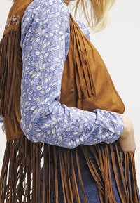 Hnědá semišová vesta s dlouhými visícími třásněmi, nošená přes modrou košili s květinovým vzorem. Vesta má hladkou texturu a neformální design.
