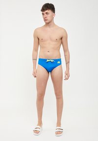 Slip da bagno blu con cordino giallo, caratterizzato da un piccolo logo sul lato sinistro. Il modello è in piedi a piedi nudi e senza maglietta.