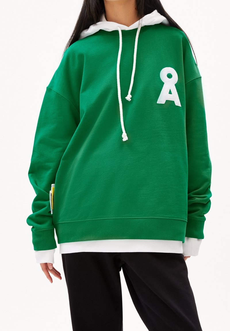 Personne portant un sweat à capuche vert avec capuche blanche et cordons, une chemise blanche superposée en dessous et un pantalon noir, debout devant un fond blanc.