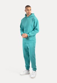 Turquoise hoodie met een voorzak en bijpassende joggers, gemaakt van zachte stof. Gecombineerd met witte sportsneakers, model staat rechtop.