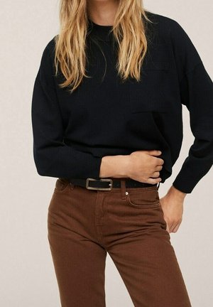 Vrouw met een zwarte trui met lange mouwen, ingestopt in een hooggetailleerde bruine broek met een zwarte riem, hand rustend op haar taille.