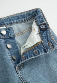 Jeans dżinsowe w niebieskim odcieniu, z kontrastującym beżowym przeszyciem, metalowymi guzikami oraz brązowym akcentem tkaniny wewnątrz paska.