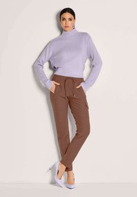 Frau in einem lavendelfarbenen Rollkragensweater, braunen Jogginghosen mit Kordelzug, lavendelfarbenen High Heels, die vor einem schlicht hellen Hintergrund steht und die Hände in den Taschen hat.
