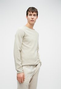 Maglione a maglia beige chiaro con collo rotondo, caratterizzato da una finitura testurizzata e maniche lunghe, abbinato a pantaloni di lino chiari.