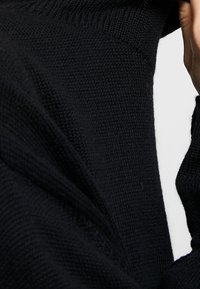 Gros plan d'une personne portant un pull en laine noir, montrant le tissu texturé et une partie de son cou et de son menton.