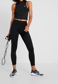 Haut court noir sans manches avec logo, associé à un legging noir taille haute. Les chaussures présentent une texture en maille avec un détail de semelle blanche.