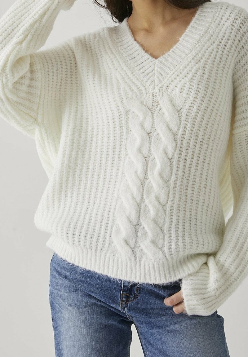 Femme portant un pull en grosse maille blanc avec un motif tressé à l'avant et un jean en denim bleu, sur un fond uni.