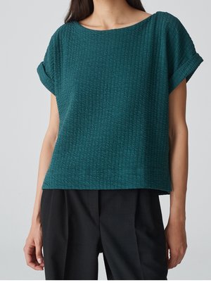 Woman wearing a teal textured short-sleeve top with rolled cuffs and loose black pants against a plain background.