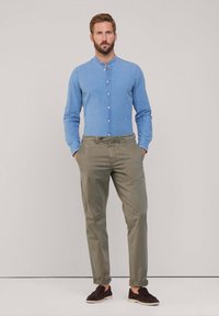 Camisa de botones azul claro con cuello mao, combinada con chinos verde oliva y zapatos oscuros sin cordones, mostrada sobre un fondo neutro.