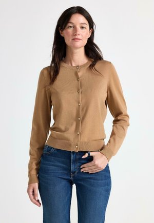 Kvinde med langt mørkt hår iført en knappet kamelfarvet cardigan og blå jeans, stående mod en ensfarvet hvid baggrund.