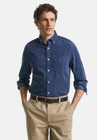 Marineblaues Langarmhemd mit Knöpfen und einer Brusttasche, kombiniert mit beigen Hosen und einem braunen Ledergürtel.