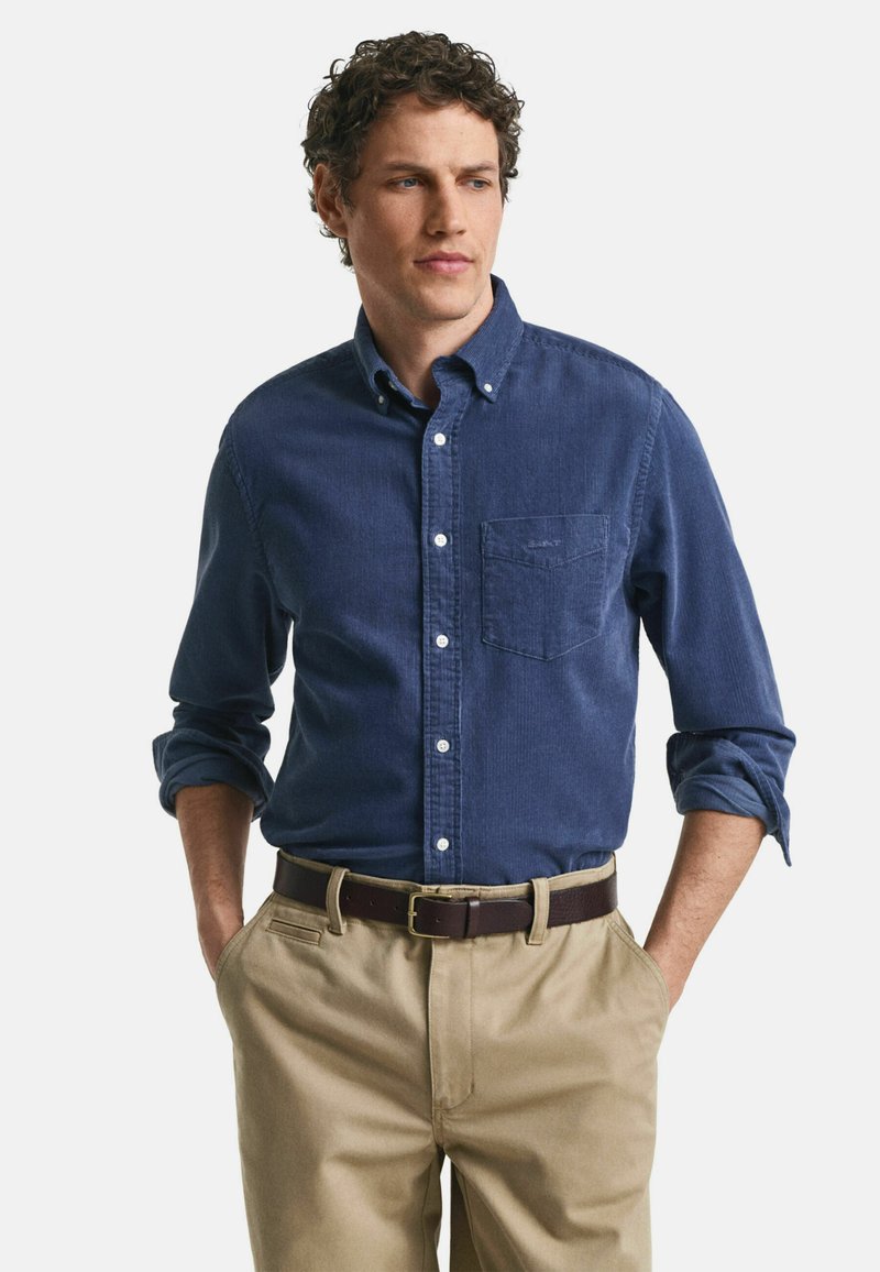 Marineblaues Langarmhemd mit Knöpfen und einer Brusttasche, kombiniert mit beigen Hosen und einem braunen Ledergürtel.