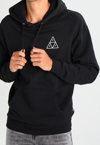 Czarna bluza z kapturem z kieszenią z przodu, regulowanymi sznurkami i białym trójkątnym logo na piersi. Materiał bawełniany, luźny krój.