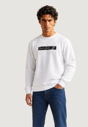 Uomo con capelli ricci che indossa una felpa bianca EA7 Emporio Armani e jeans blu, in piedi con una mano in tasca su uno sfondo semplice.