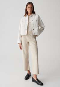 Giacca di jeans bianca con due tasche frontali, abbinata a jeans beige a gamba dritta. Mocassini neri completano l'outfit. Design pulito e minimale.