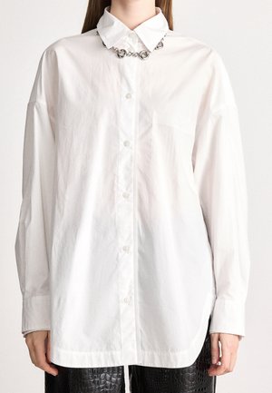 Persoon die een oversized wit overhemd met puntige kraag en kettinkdetail draagt, gecombineerd met zwarte gestructureerde broek.