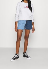 Donna che indossa pantaloni corti in denim bicolore, una felpa grigia chiara con logo e scarpe da ginnastica alte nere, in piedi su uno sfondo neutro.