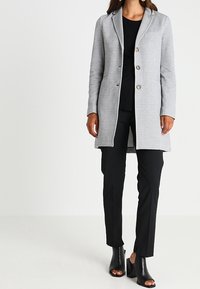 Veste longue gris clair avec un col cranté, trois boutons et une poche poitrine, associée à un pantalon noir ajusté et des talons ouverts noirs.