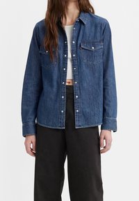 Donna che indossa una camicia di jeans blu aperta sopra un crop top bianco e pantaloni larghi neri, con lunghi capelli castani visibili.
