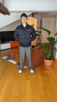 Casaco preto acolchoado com fecho de correr à frente e punhos canelados, usado por cima de calças cinzentas com padrão, em cima de chão de madeira com plantas de interior por perto.