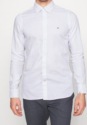 Muž v bílé košili s malými modrými puntíky a tmavě šedých kalhotách, stojící před jednobarevným pozadím.