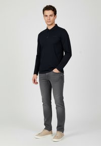 Czarna koszulka polo z długim rękawem i plisą zapinaną na guziki, zestawiona z szarymi jeansami o dopasowanym kroju oraz beżowymi sneakersami, na neutralnym tle.