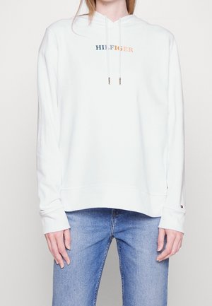 Person trägt weißen Hoodie mit mehrfarbigem "HILFIGER" Schriftzug und hellblaue Jeans, steht vor einfachem hellem Hintergrund.