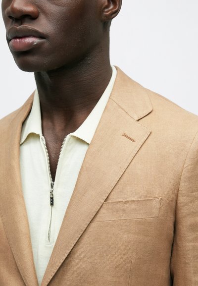 Κοντινό πλάνο ενός ατόμου που φοράει ένα μπεζ σακάκι πάνω από ένα λευκό πόλο με φερμουάρ, με φόντο ένα απλό υπόβαθρο, εμφανίζοντας τον λαιμό και το κάτω μέρος του προσώπου.