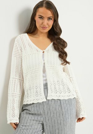 Mujer con cabello largo y ondulado, vestida con un cárdigan blanco de punto sobre una camiseta blanca y pantalones grises con rayas verticales, de pie frente a un fondo claro.