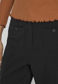 Bruine ribgebreide top met lange mouwen en geribbelde zoom, gecombineerd met zwarte op maat gemaakte broek met twee voorzakken en een knopenaccent.