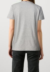 Vrouw in een effen lichtgrijze korte mouwen t-shirt en zwarte broek, gezien vanuit de achterkant tegen een witte achtergrond.