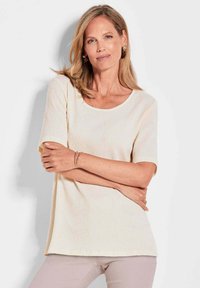 Crème, geribbelde top met korte mouwen en een ronde halslijn. Zachte textuur en een relaxte pasvorm, gecombineerd met lichte broek en geaccessoriseerd met een armband.