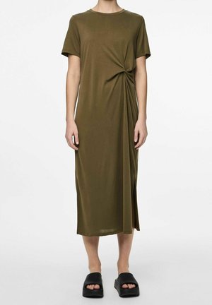 Kvinde iført en olivengrøn midi-kjole med korte ærmer og sidesløjfedetalje, kombineret med sorte platform-slider-sandaler, stående på hvid baggrund.