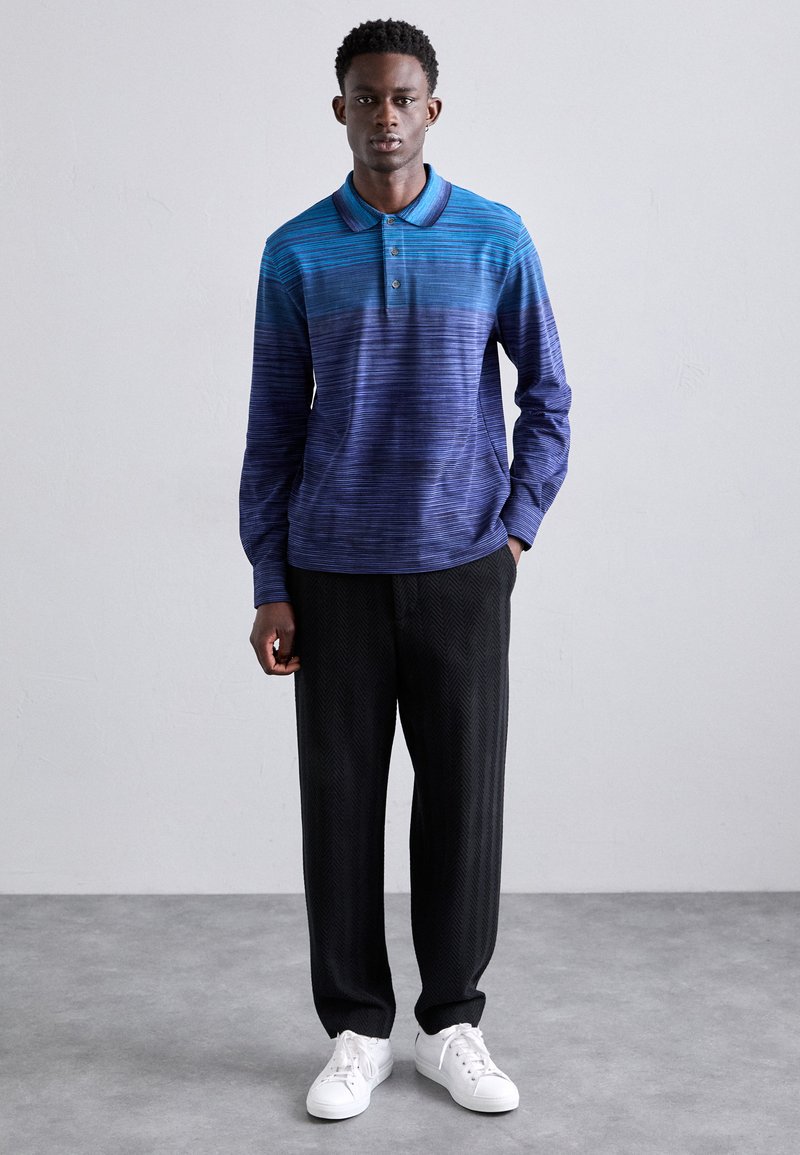 Langærmet polo i gradient blå nuancer, med krave, knaplukning og parret med sorte herringbone bukser og hvide sneakers.