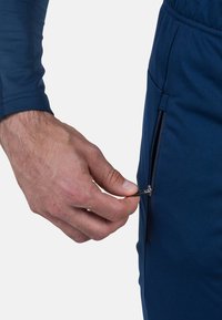 Marjapuna urheiluhousut, joissa on sileä pinta ja sivutasku vetoketjulla. Kädessä pidetään vetoketjun vedintä, mikä korostaa käytännöllistä muotoilua.