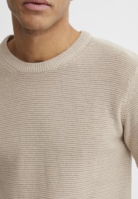 Bež pleteni pulover s okruglim izrezom, s teksturiranim rebrastim uzorkom, napravljen od mekanog, prozračnog materijala.