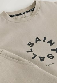 Bež jopica z okroglim izrezom, okrašena s krožnim črnim grafičnim napisom "ALLSAINTS" in "LONDON" na dnu. Bombažno blago, gladka tekstura.